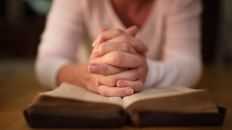A woman's hands clasped in prayer rest on a Bible