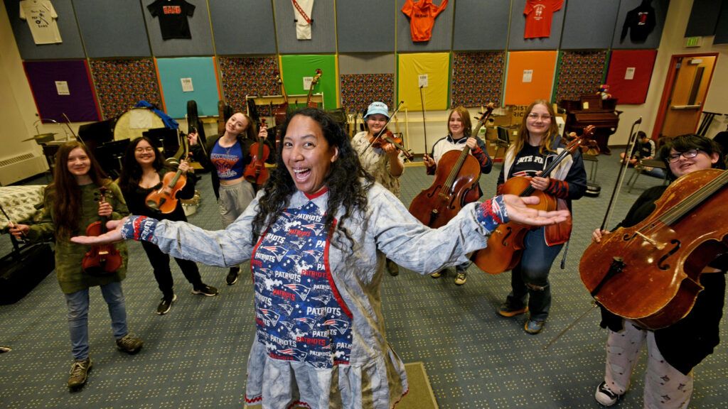 A smiling music teacher stands in front of her students with her arms raised up