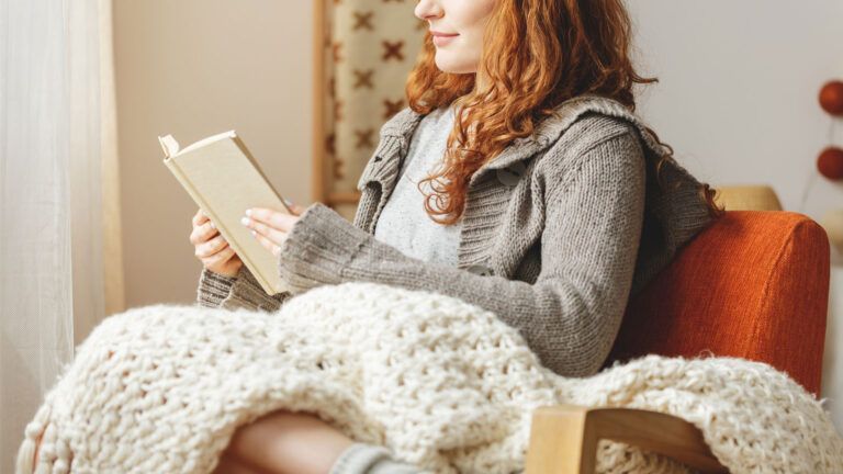 Woman reading in a cozy chair with a large knitted blanket.